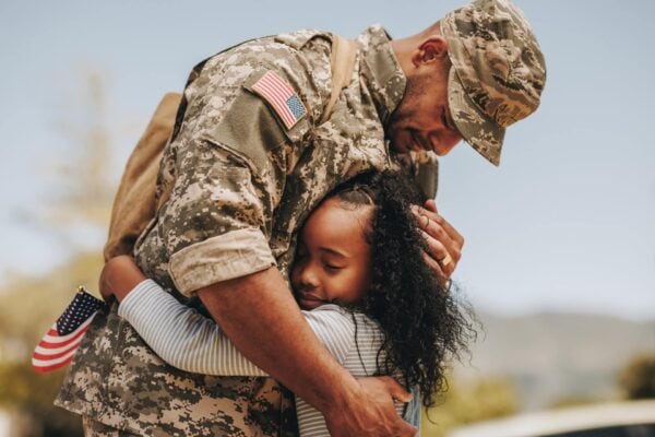 A soldier in camouflage uniform hugs a young girl holding a small American flag outdoors.