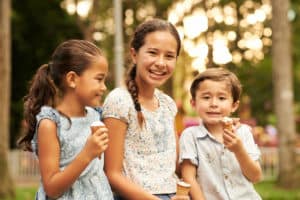 Three children sit outdoors eating ice cream cones, smiling and appearing happy, with trees and blurred background visible.