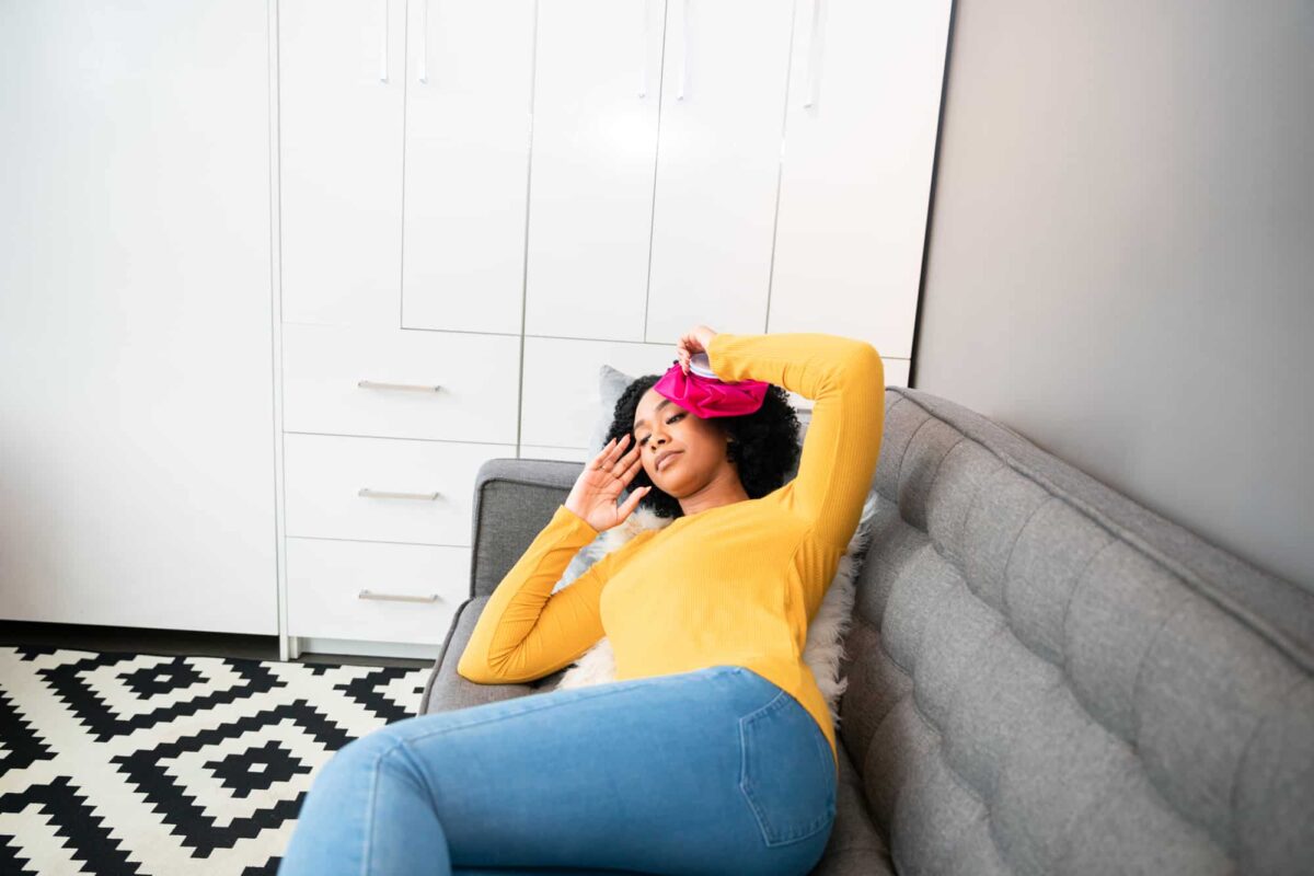 Woman lying on a gray couch holding a pink ice pack on her forehead, appearing unwell, in a modern living room with white cabinets.