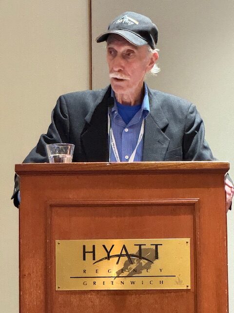 An older man in a black cap and blazer speaks at a podium with a "Hyatt Regency Greenwich" sign and a glass of water.