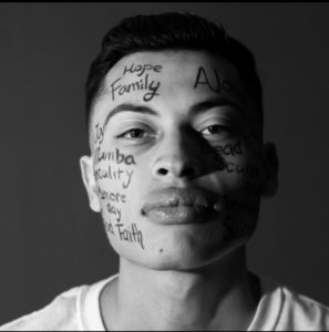 A man gazes forward with words such as "Hope," "Family," and "Faith" written in black marker across his face; photo is in black and white.
