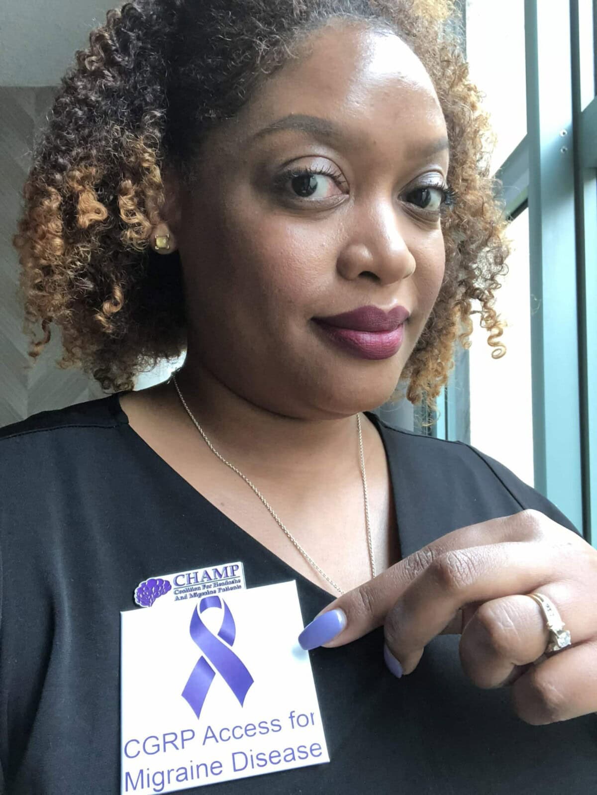 A woman with curly hair holds a card reading "CGRP Access for Migraine Disease" with a purple ribbon and a CHAMP sticker, standing by a window.
