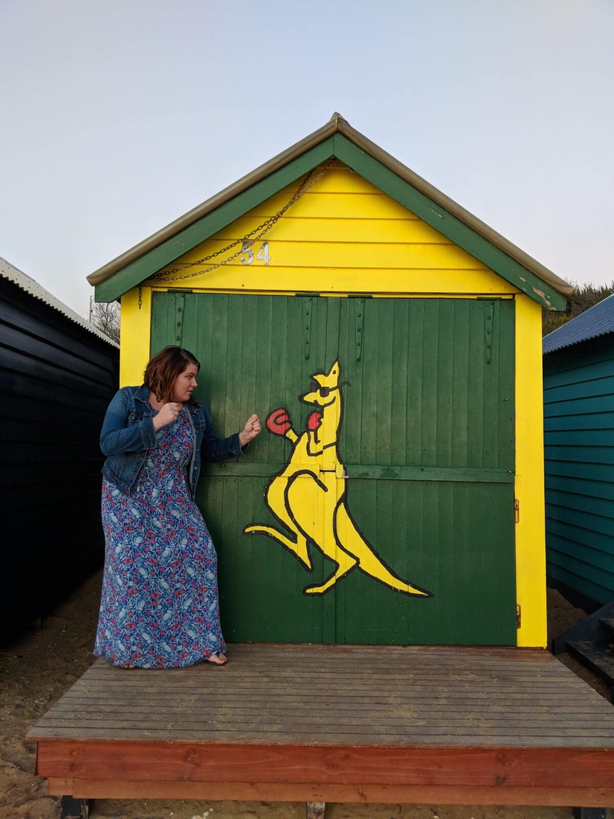 A woman in a long dress playfully poses as if boxing with a yellow kangaroo mural painted on a green and yellow shed.