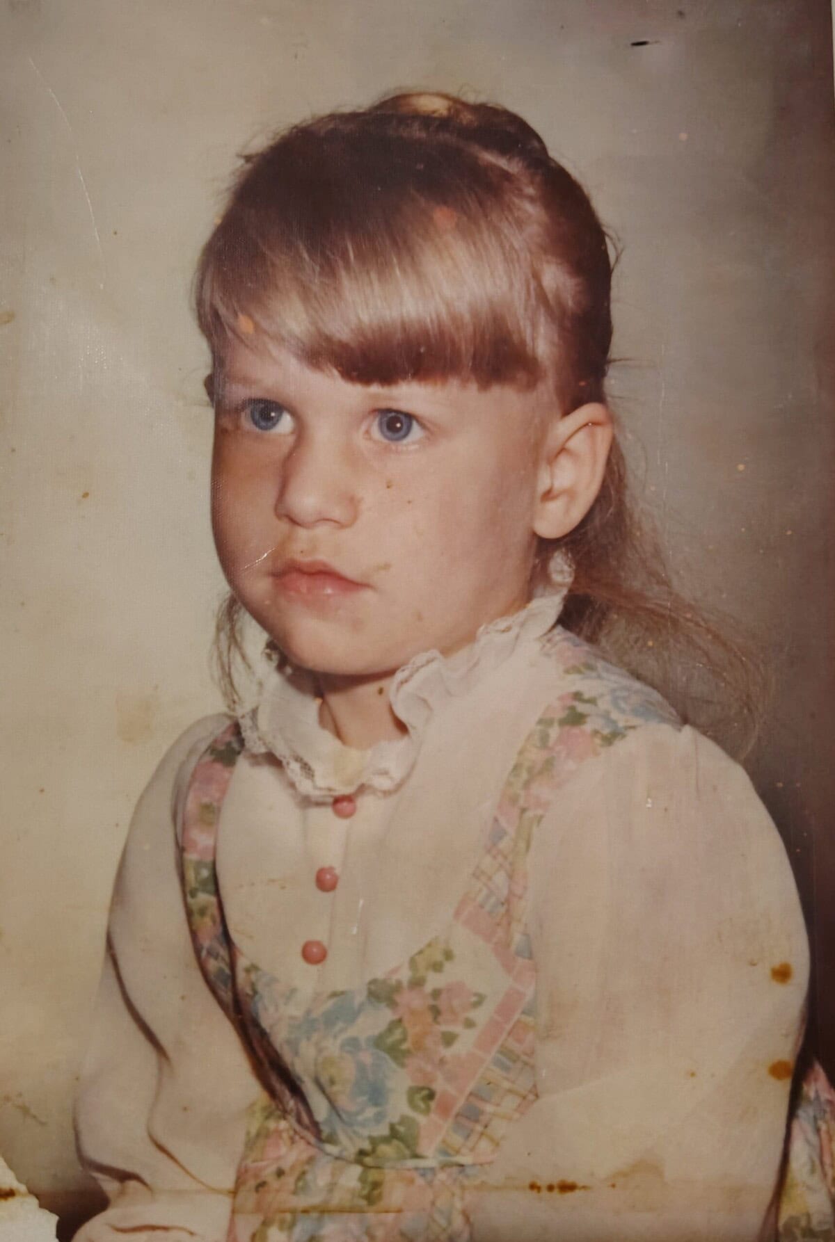 A young girl with light brown hair and blue eyes sits for a portrait, wearing a floral dress with a white blouse and pink buttons, against a plain background.