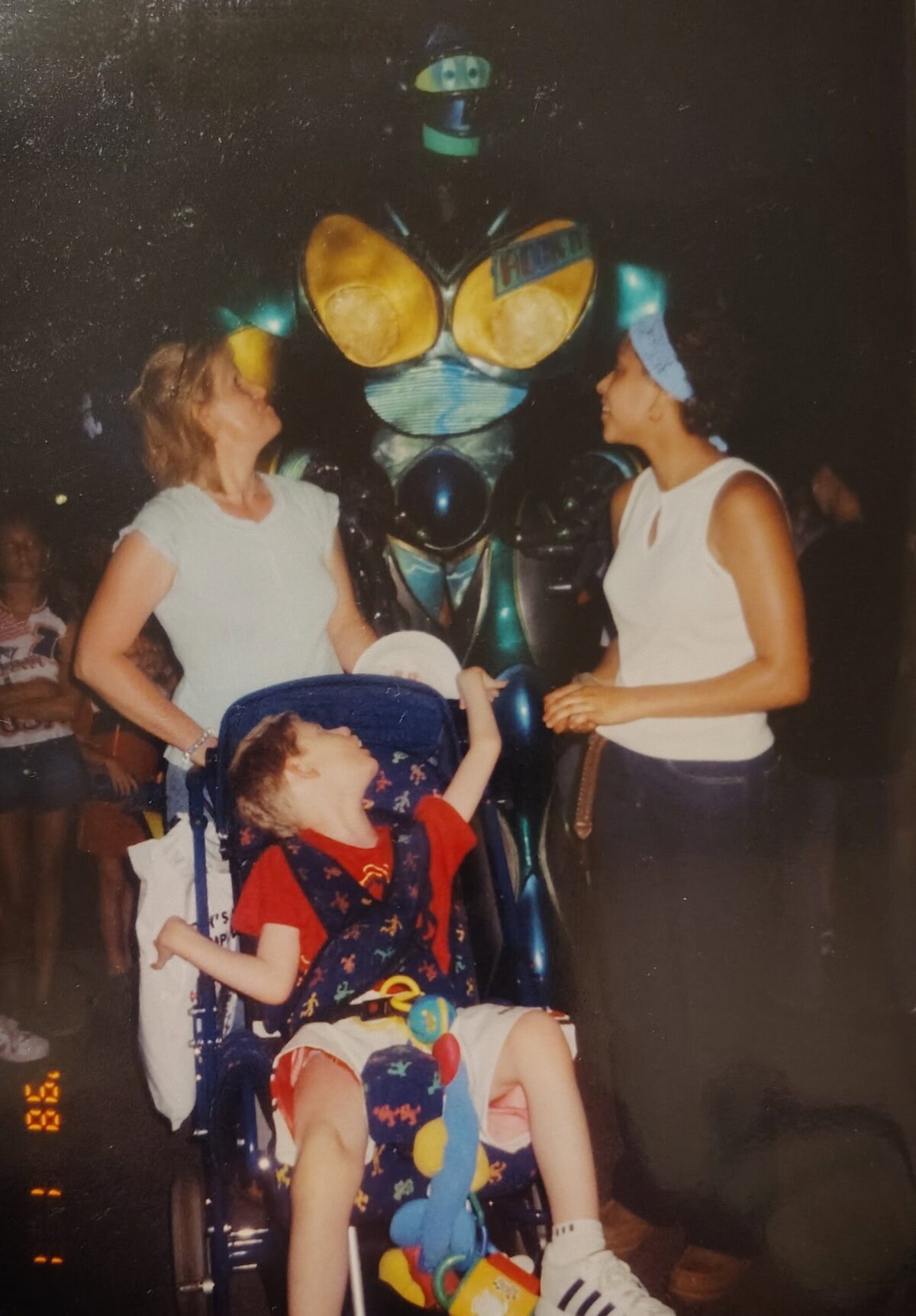 A child in a stroller and two women stand in front of a tall person in a robot costume at an outdoor event at night.