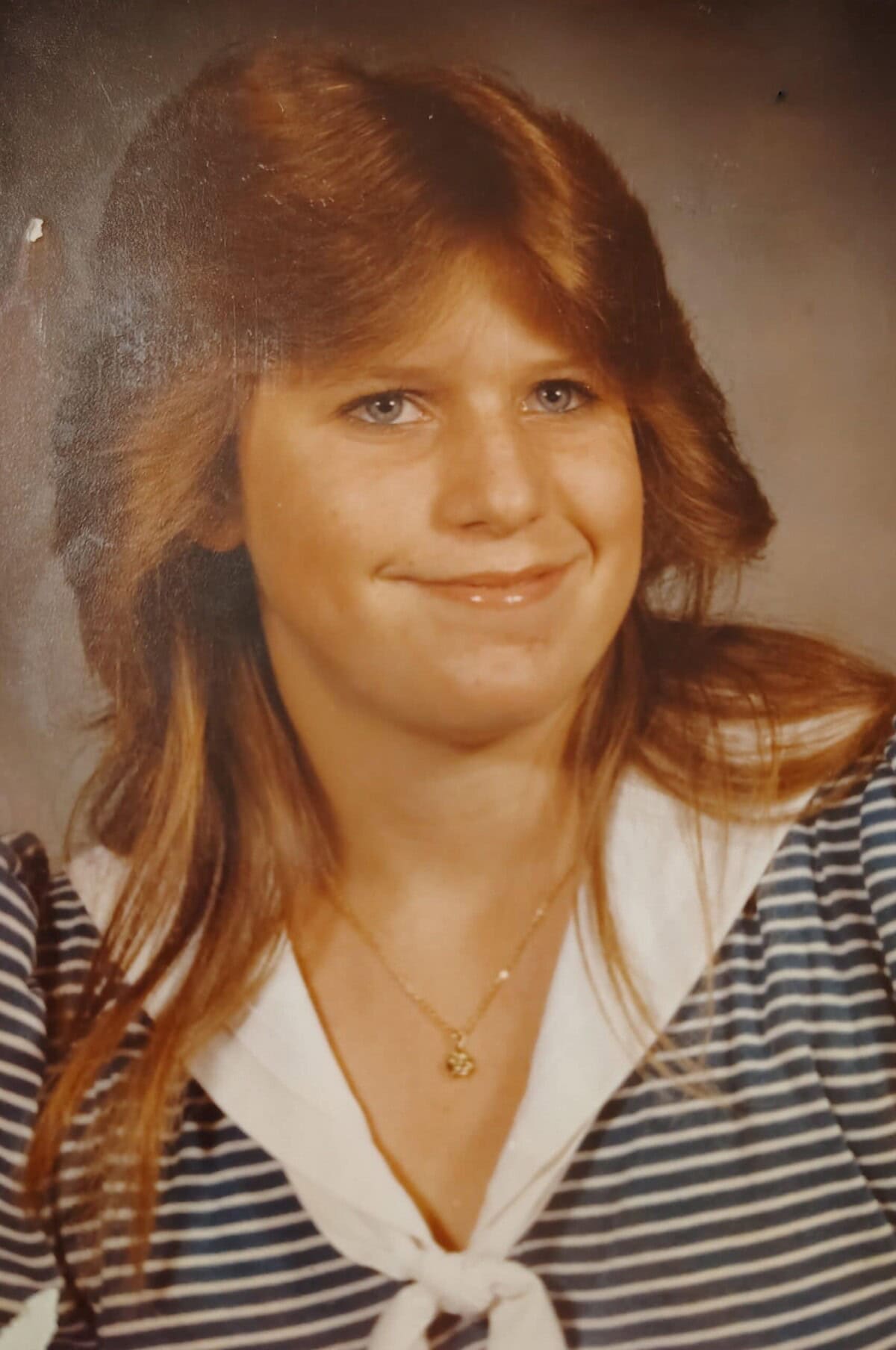 A young woman with long, light brown hair and blue eyes smiles at the camera. She is wearing a striped shirt with a white collar and a gold necklace.