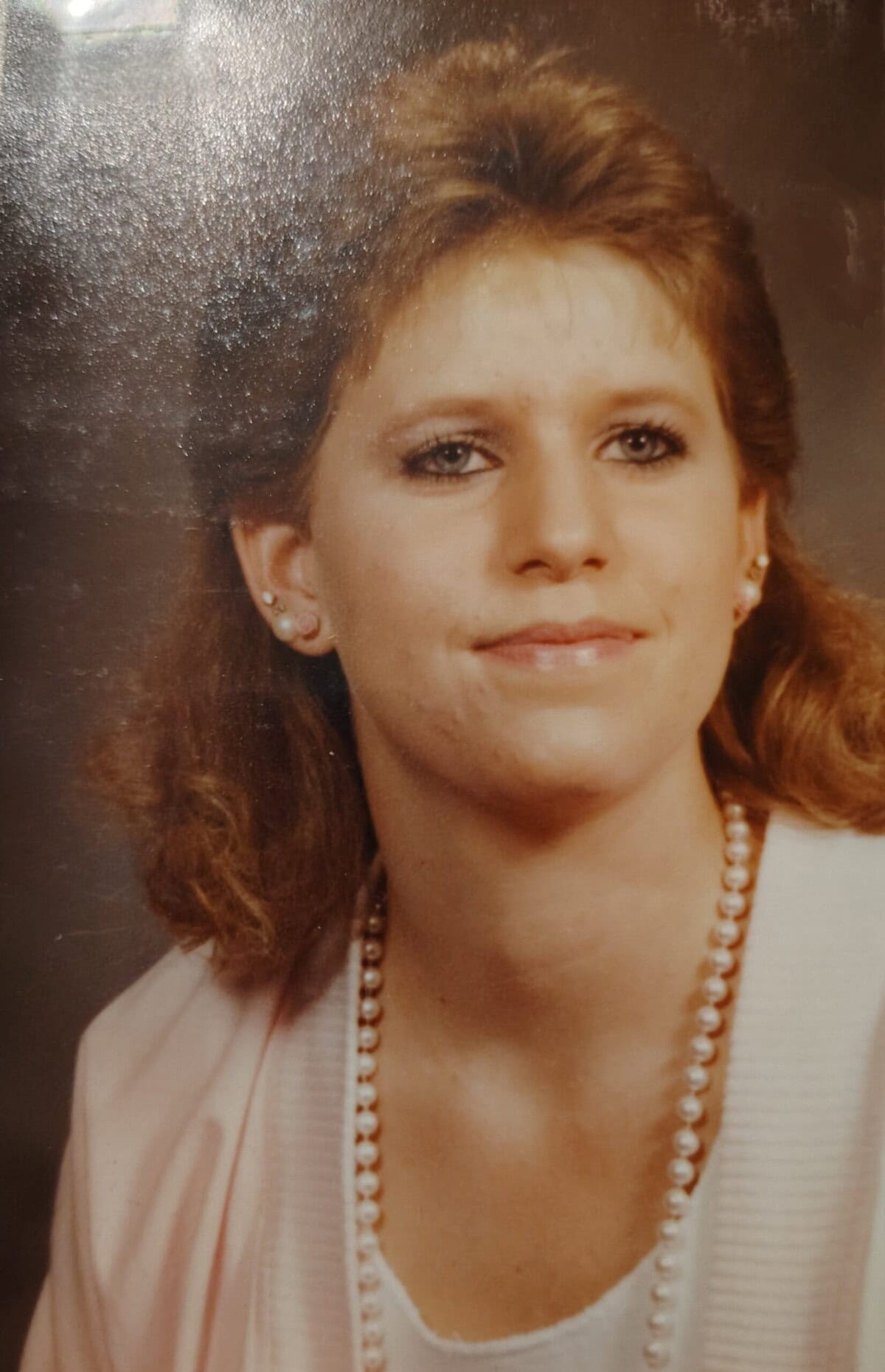 A woman with shoulder-length light brown hair, wearing a light pink blazer, white top, pearl necklace, and small earrings, poses in front of a plain brown background.