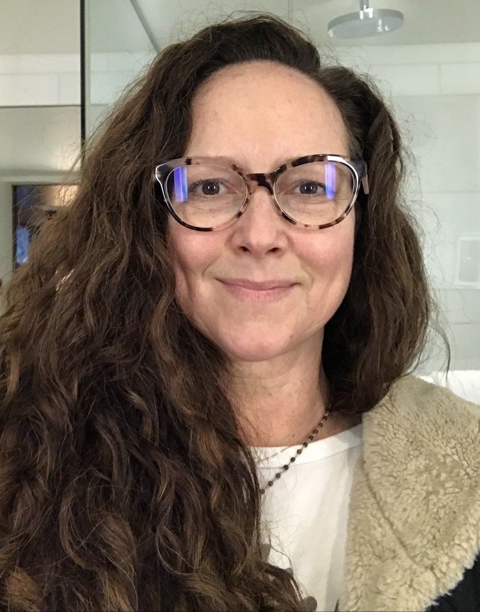 Woman with long curly brown hair and glasses wearing a white top and a light-colored jacket, standing indoors in front of a mirror.
