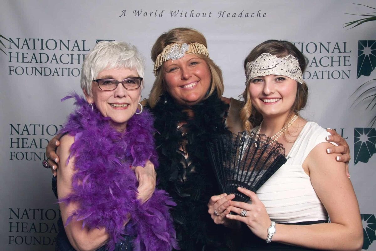 Three women in 1920s-style outfits and feather boas pose together, smiling, in front of a National Headache Foundation backdrop.