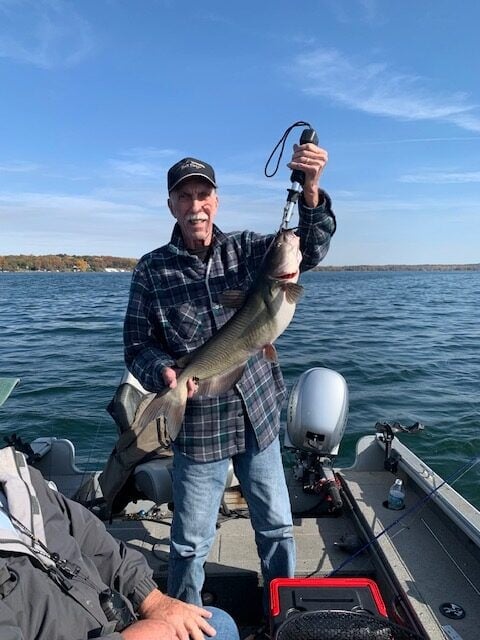 An older man stands on a boat holding a large fish with a gripping tool, smiling at the camera. The boat is on a lake under a clear blue sky.