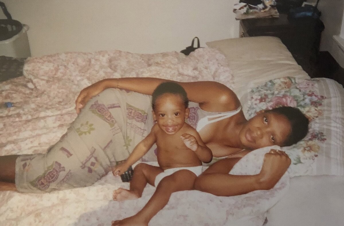 A woman lies on a bed next to a smiling baby, both facing the camera. The baby is sitting up, and the woman is resting on her side with one arm under her head.