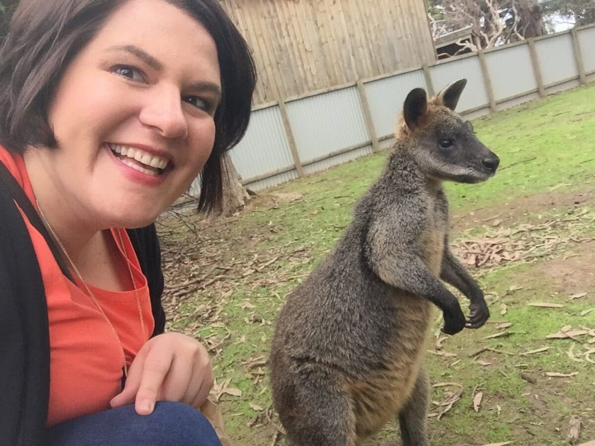 A woman smiles and poses for a selfie next to a wallaby in an outdoor enclosure with a wooden fence in the background.