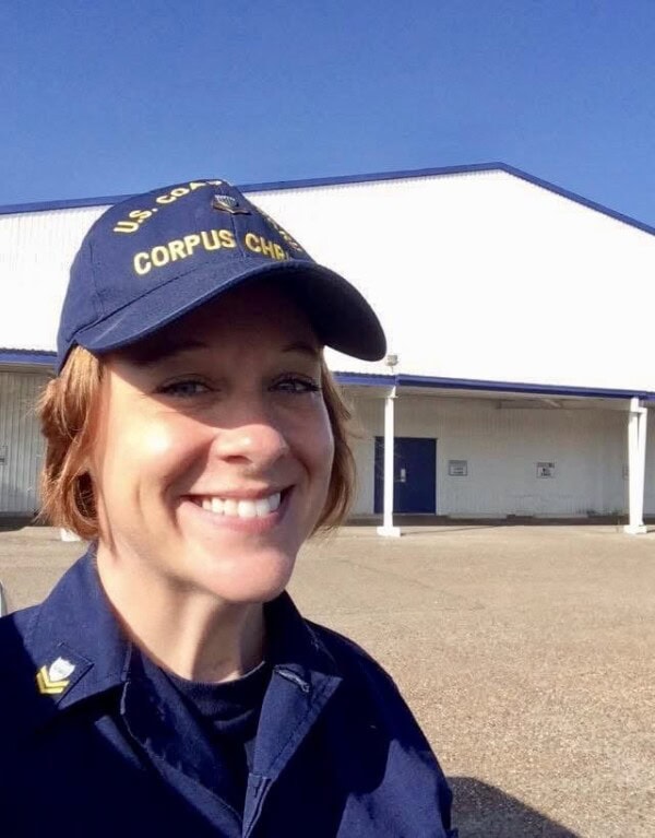 A person in a U.S. Coast Guard uniform and cap stands outside, smiling, with a large white building in the background.