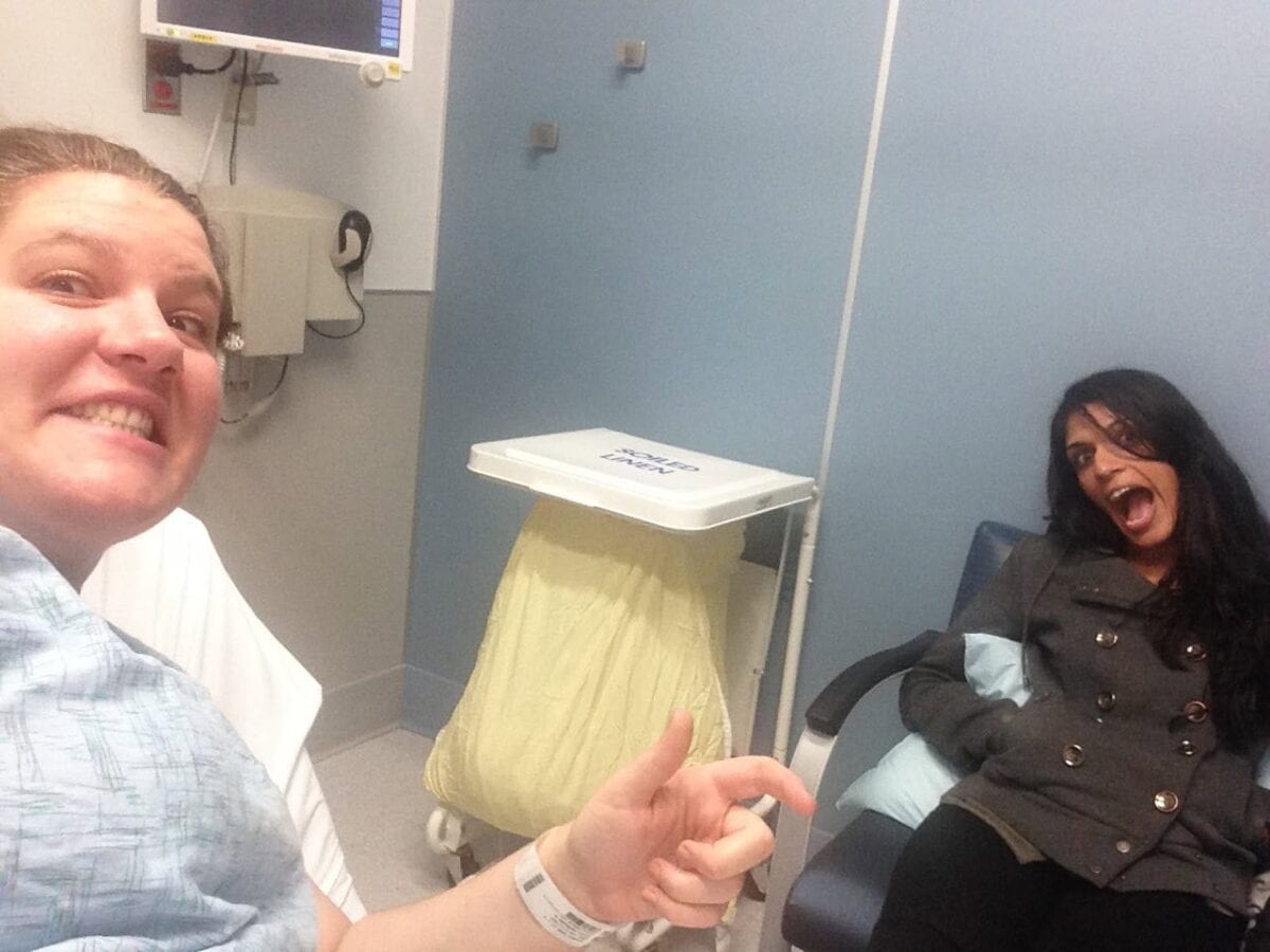 Two women are in a hospital room, one in a hospital bed wearing a gown and wristband, smiling, and pointing at the other woman who is sitting in a chair, also smiling.