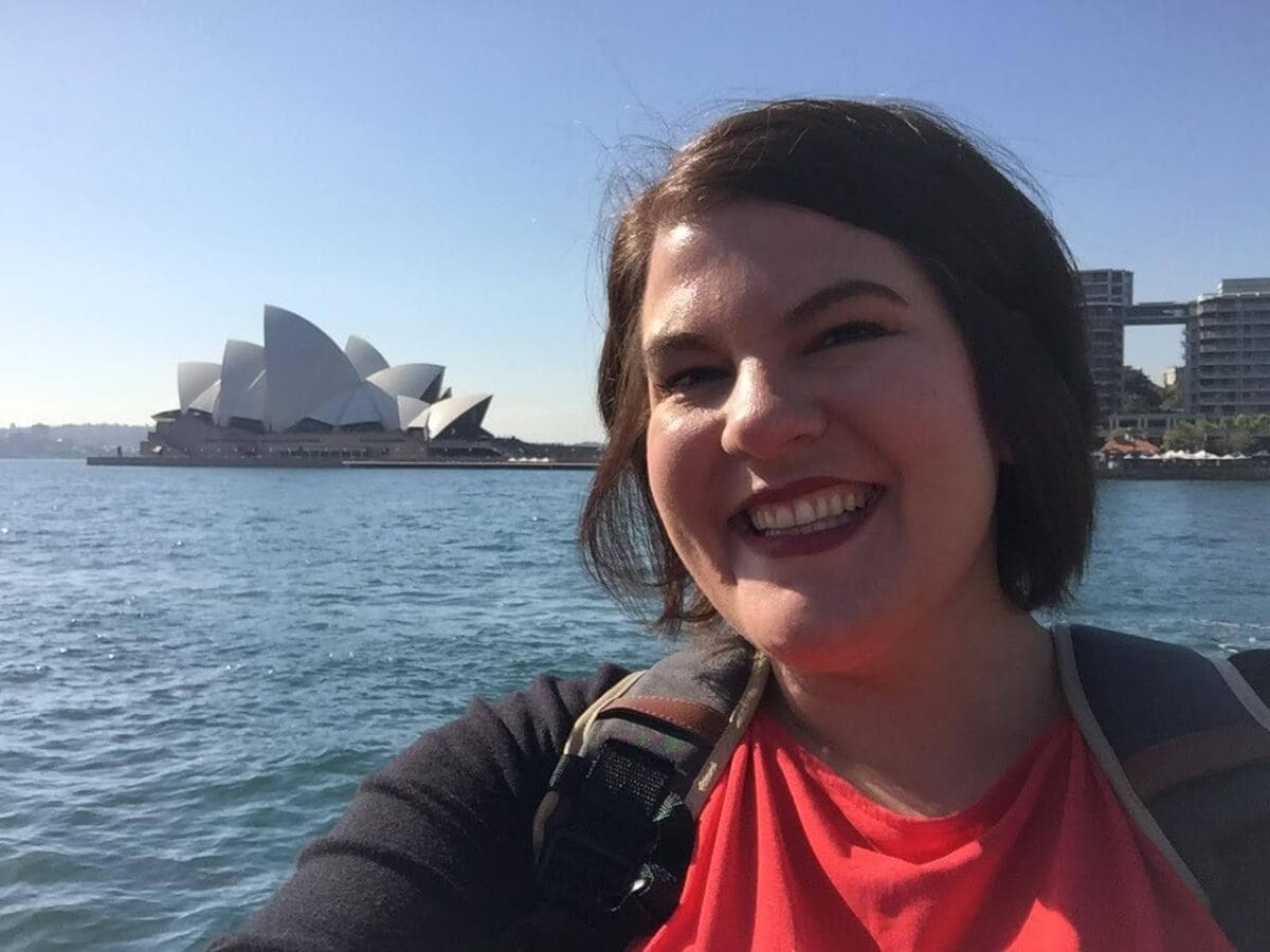 A woman takes a selfie near the water with the Sydney Opera House and city buildings in the background on a sunny day.
