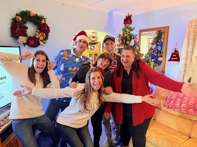 Six adults pose together in a living room decorated for Christmas, smiling and making playful gestures in front of a tree and festive decorations.