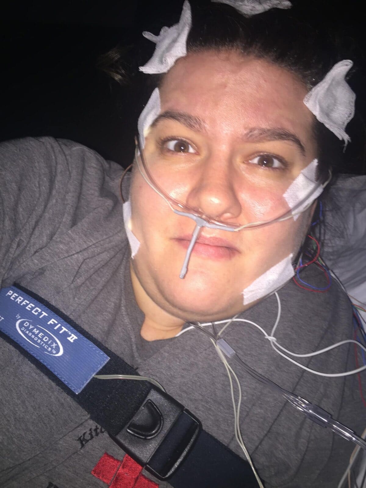 Person lying down with nasal cannula, electrodes, and adhesive pads attached to their face and head, wearing a grey shirt and a seatbelt labeled "Perfect Fit 2.
