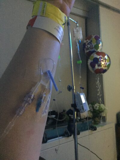 A close-up of a person’s arm with a hospital IV inserted; medical bracelets are visible, and "Get Well" balloons and equipment are in the background.