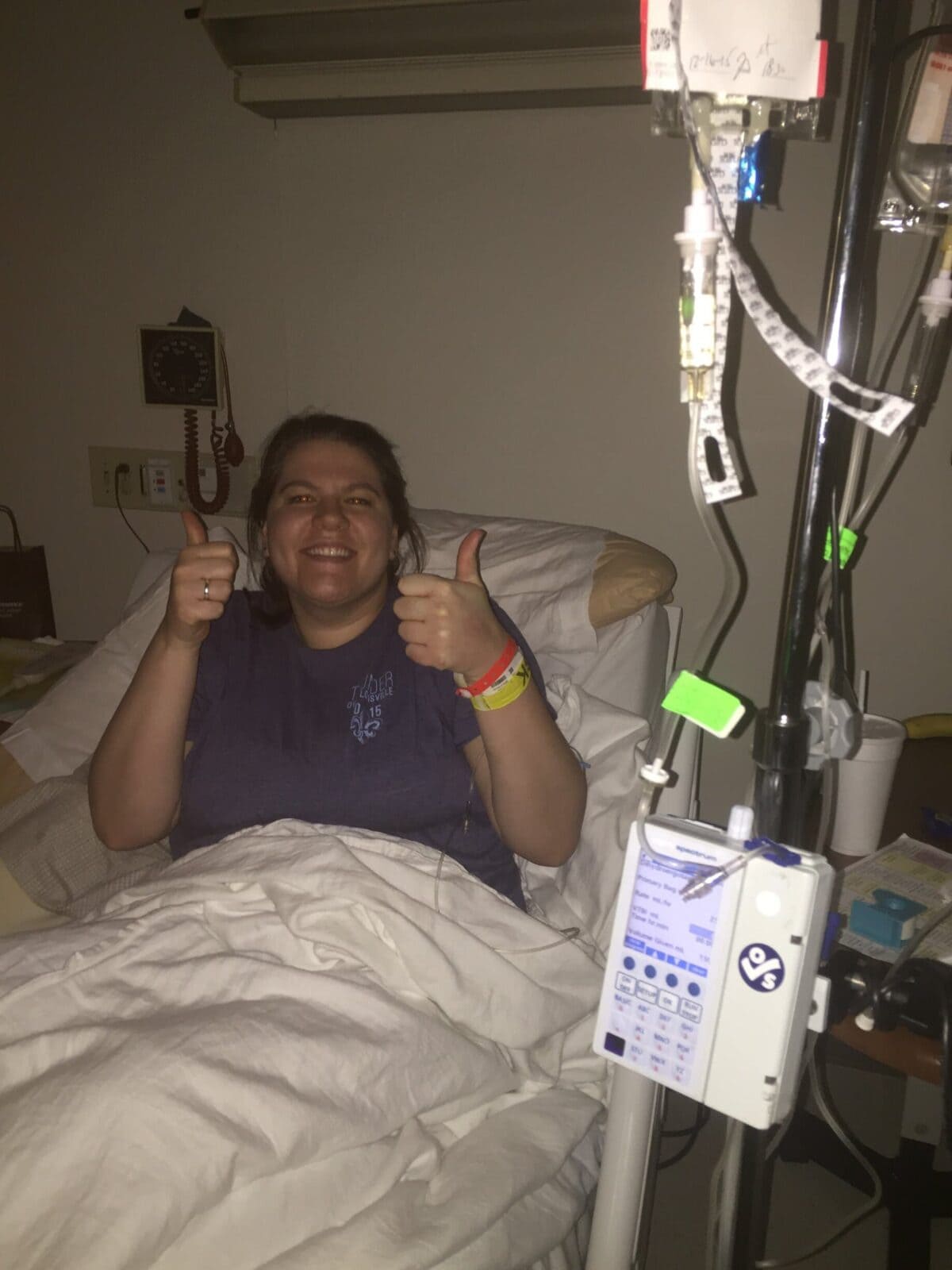 A woman in a hospital bed smiles and gives two thumbs up, with IV equipment and monitors beside her.