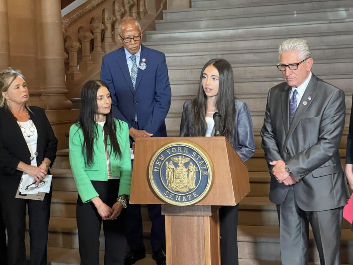 A young woman speaks at a New York State Senate podium, surrounded by four adults standing on a staircase in a formal setting.