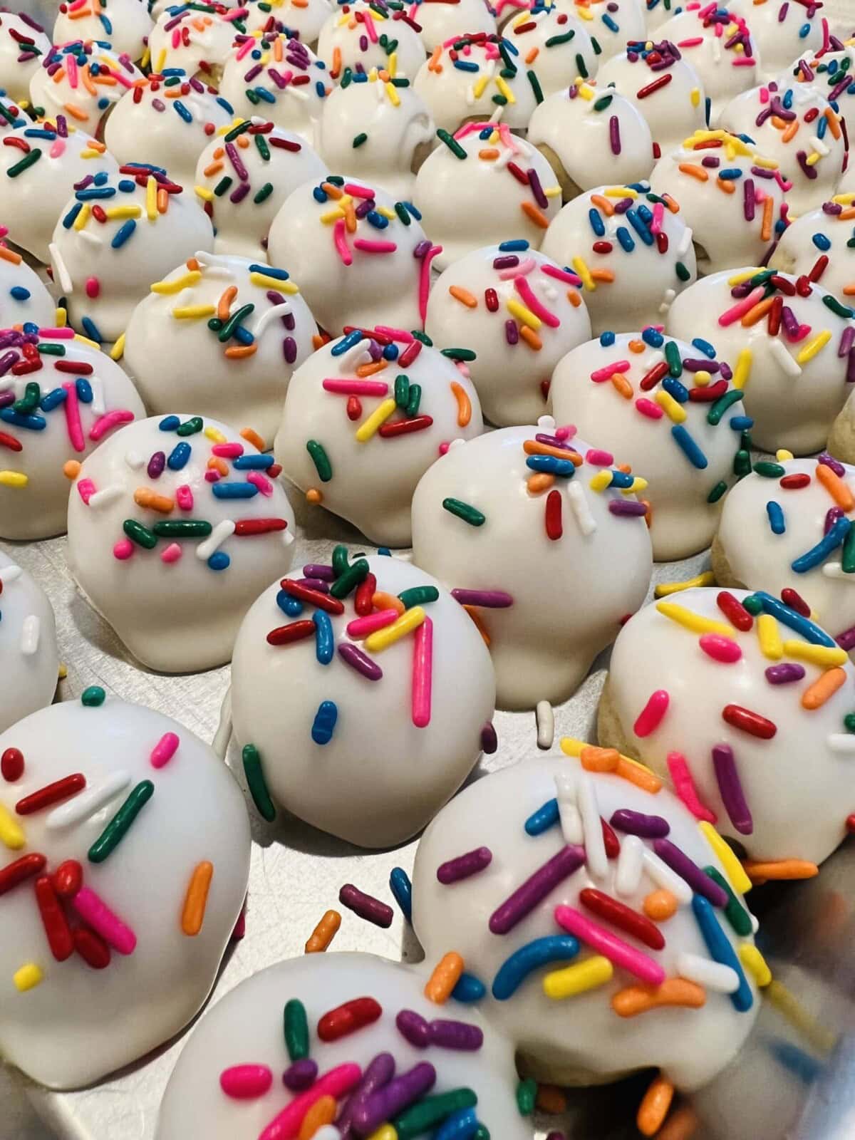Rows of round white cake balls coated in icing and topped with colorful sprinkles arranged on a baking sheet.