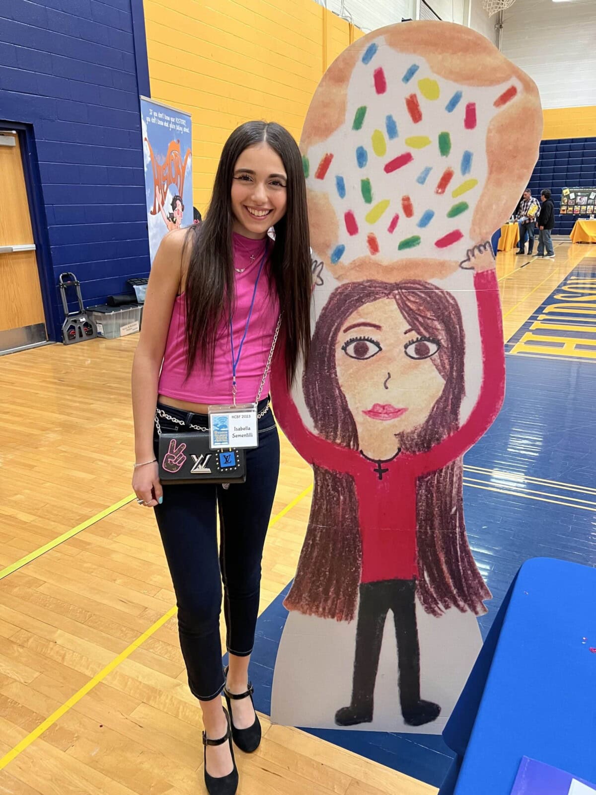 A young woman stands indoors next to a large cutout of a drawing of a girl holding a donut with sprinkles. Both are smiling.