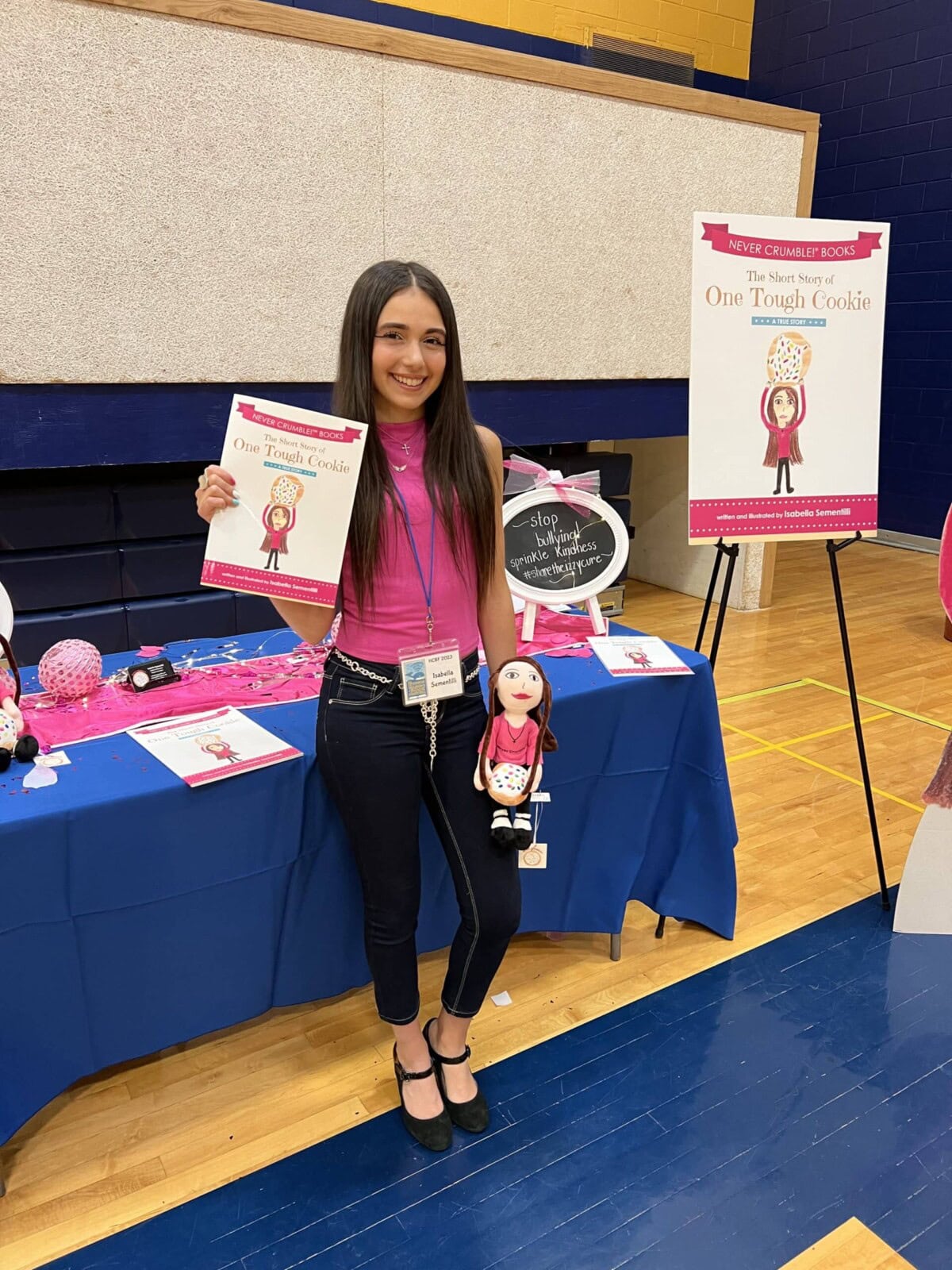 A woman stands indoors holding a children's book and a matching doll at a display table featuring promotional materials for "One Tough Cookie.