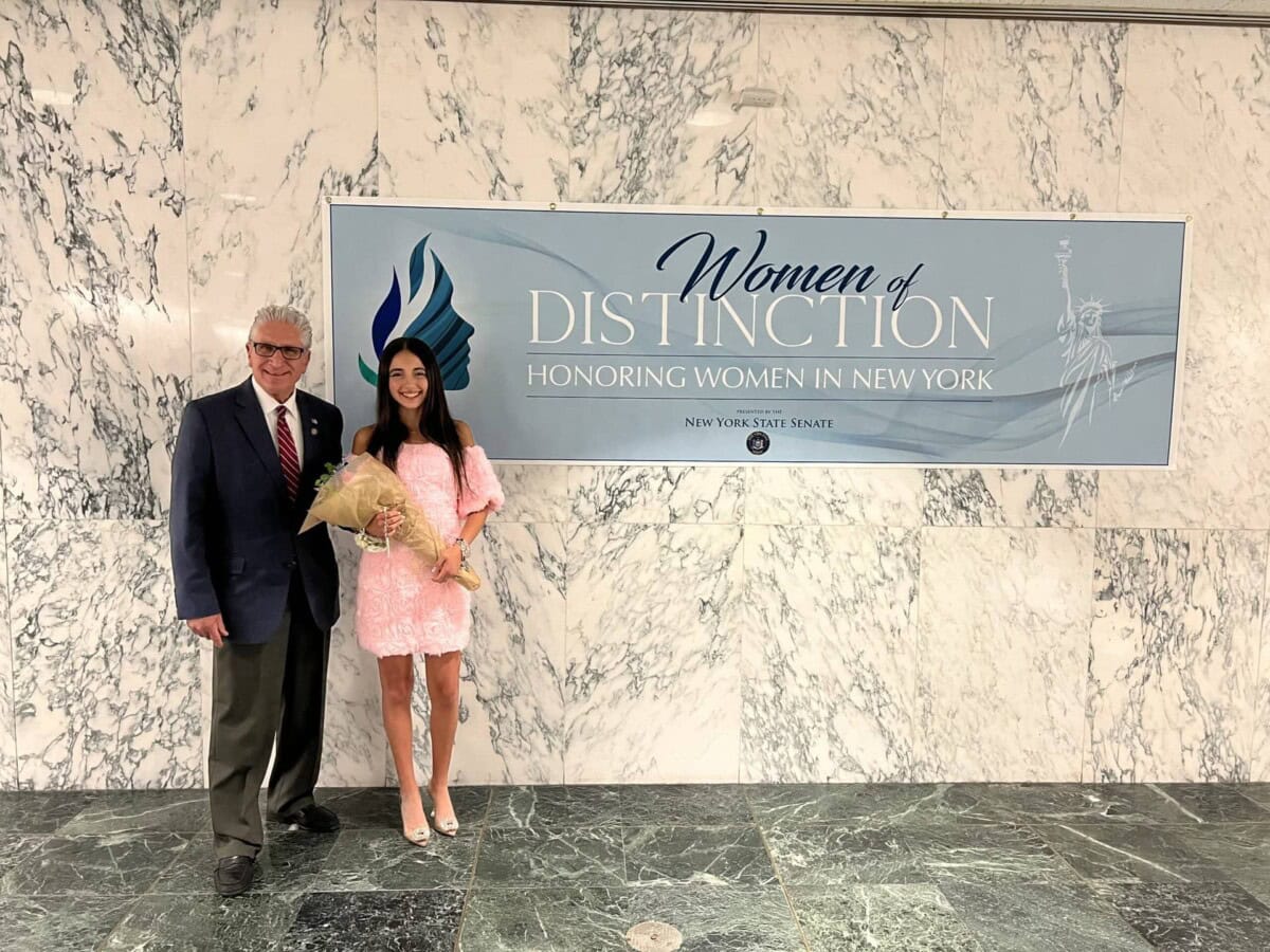 A man and a woman holding flowers stand in front of a marble wall with a sign reading "Women of Distinction: Honoring Women in New York.