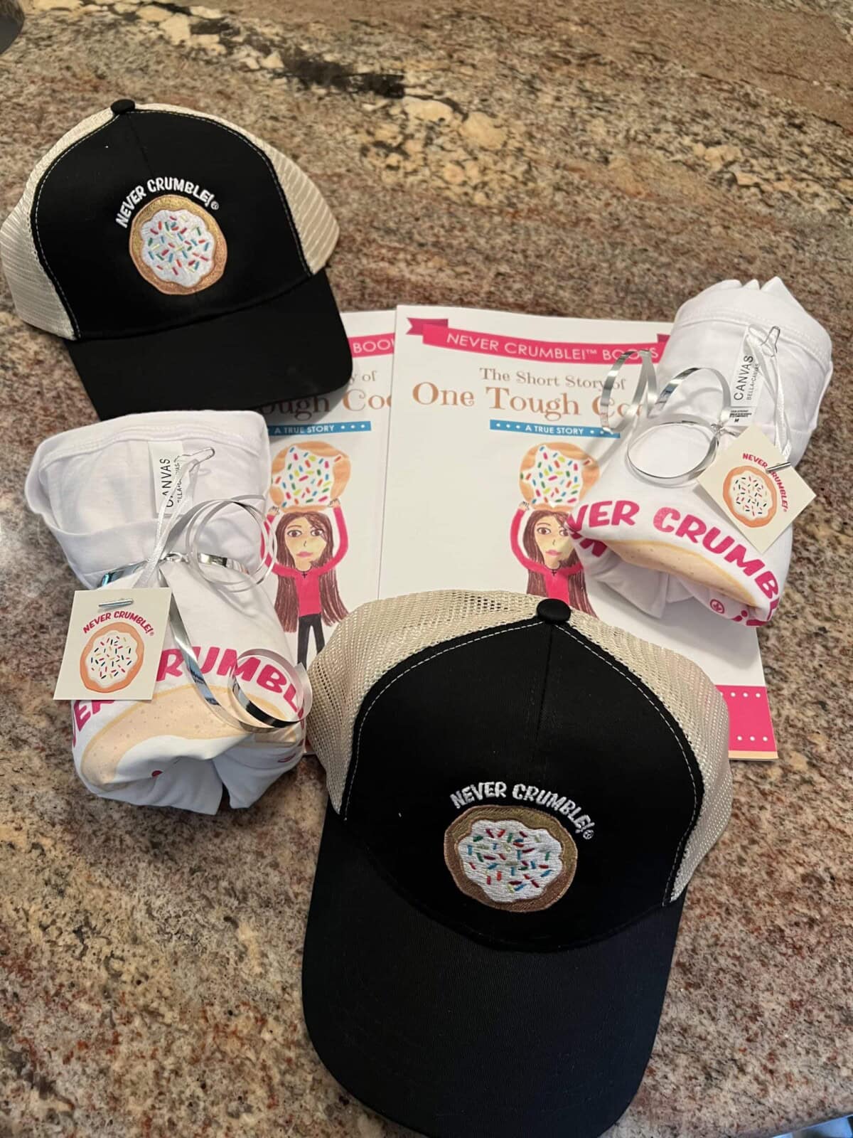 Two trucker hats with a donut logo and "Never Crumble" text, two rolled-up T-shirts, and two books titled "One Tough Cookie" displayed on a granite countertop.
