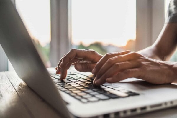 Close-up of a person's hands typing on a laptop keyboard near a window with sunlight in the background.