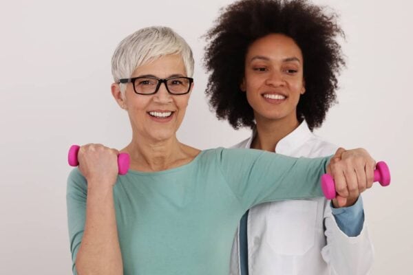 Older woman in glasses exercises with pink dumbbells while being assisted by a younger woman in a white coat, likely a healthcare or fitness professional, against a plain background.