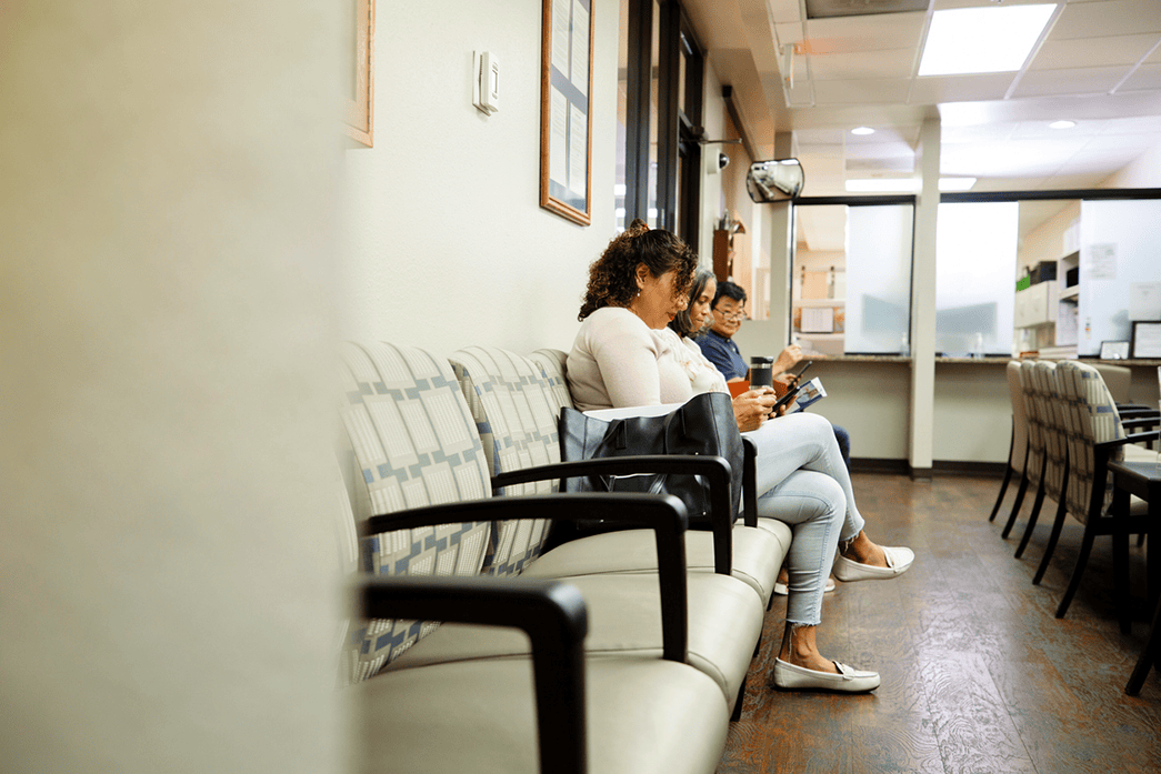 Three people sit on chairs in a waiting room, looking at their phones and papers. The room is well-lit, with empty seats and a reception area in the background.
