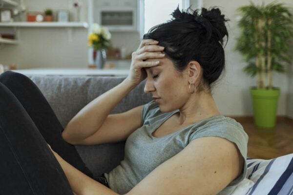 A woman sits on a couch with her eyes closed and one hand on her forehead, appearing stressed or tired.