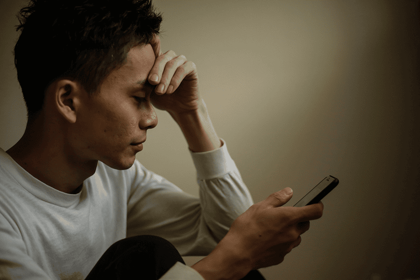 A man in a white shirt sits with his hand on his forehead, looking down at a smartphone with a concerned expression.