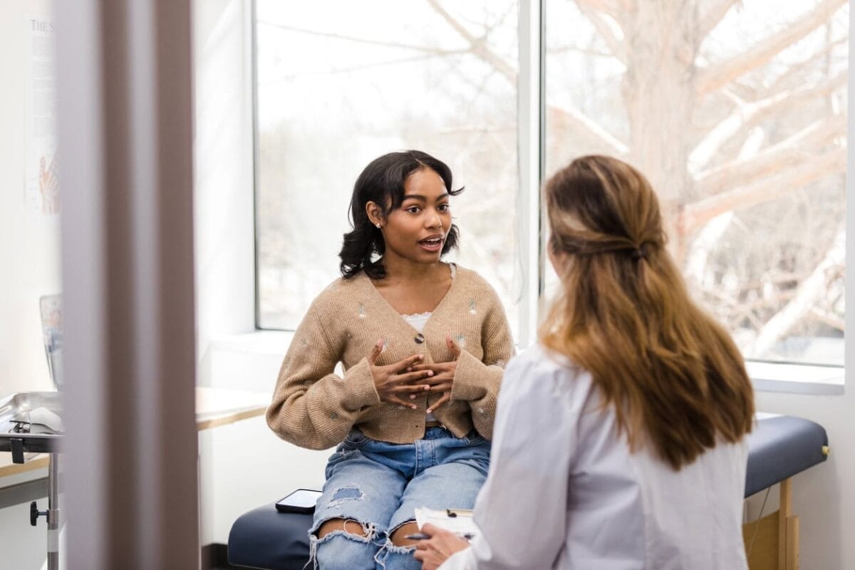 A woman sits on an exam table speaking to a healthcare professional in a white coat in a medical office with large windows.
