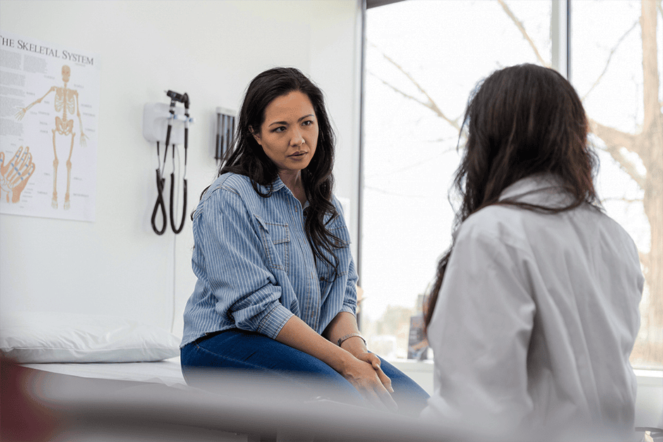 A woman sits on an exam table talking to a healthcare professional in a medical office with anatomical posters on the wall.