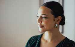 A woman with dark hair in a low bun, wearing silver hoop earrings and a dark top, looks to the side with a neutral expression.