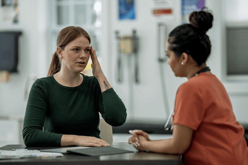 A woman in a green shirt describes symptoms to a healthcare professional in orange scrubs during a medical consultation at a clinic.
