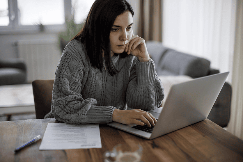 A woman in a gray sweater sits at a table using a laptop, with documents and a pen on the table in front of her.