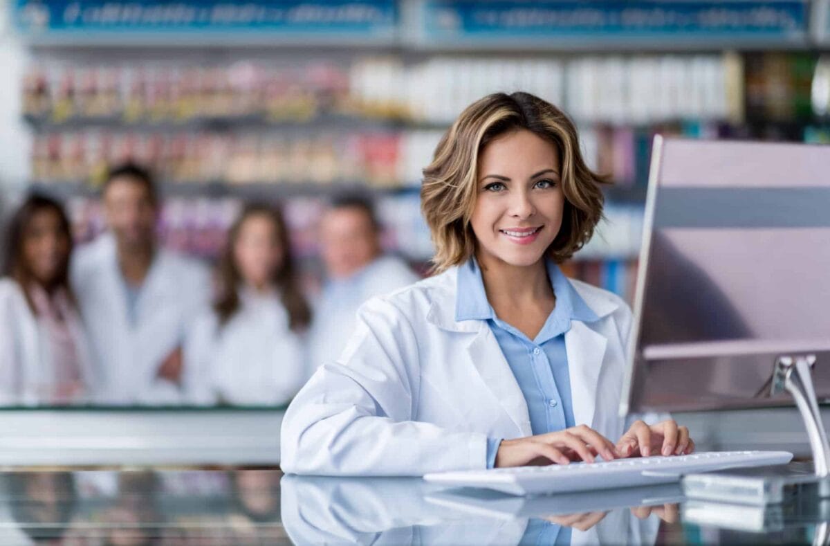 A pharmacist in a white coat types on a computer at a pharmacy counter, with four colleagues standing in the blurred background among shelves of products.