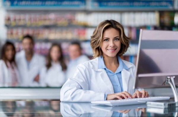 A pharmacist in a white coat types on a computer at a pharmacy counter, with four colleagues standing in the blurred background among shelves of products.