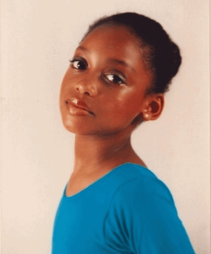 A young girl with dark hair pulled back is wearing a blue top and looking at the camera against a plain light background.