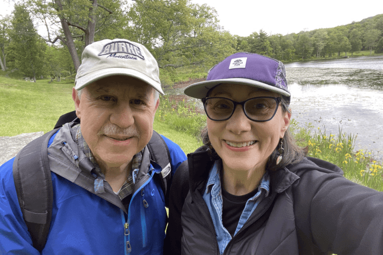 Two adults wearing hats and jackets smile for a selfie outdoors near a pond, with green trees and grass in the background.