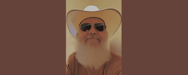 A man with a white beard wearing a white cowboy hat, sunglasses, and a brown shirt is facing the camera and smiling.