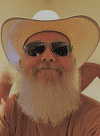 A man with a long white beard wears a cowboy hat and sunglasses, smiling at the camera indoors.