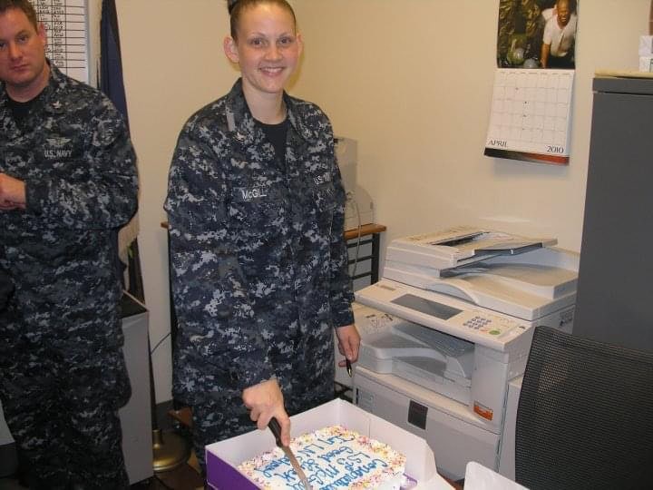 A person in a Navy uniform cuts a cake in an office setting, with a printer and a calendar on the wall in the background.