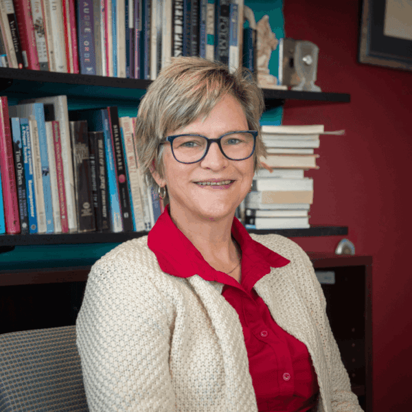 A woman with short blonde hair and glasses, wearing a red shirt and cream cardigan, sits and smiles in front of a bookshelf filled with books.
