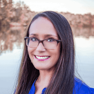 Woman with long straight brown hair and glasses, wearing a blue top, smiling in front of a calm lake with trees in the background.