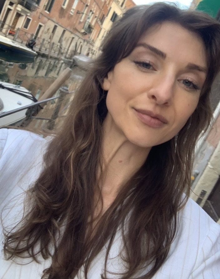 A woman with long brown hair takes a selfie near a canal with boats and old buildings in the background.