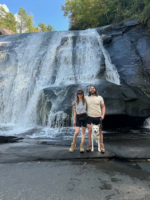 A woman, a man, and a white dog stand in front of a large waterfall with trees at the top of the falls.